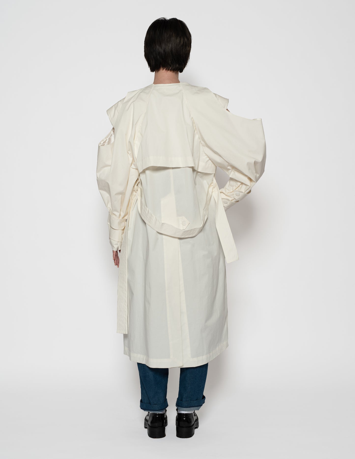 Back view of a long white trench coat with pleated detail and detachable sleeve overlay.