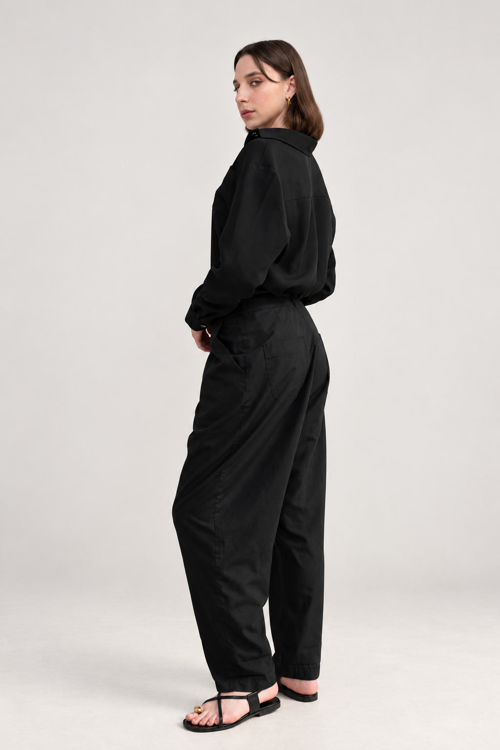 “Back three-quarter view of Casa Como black relaxed trousers styled with a black long-sleeve shirt, showing the rear pockets and relaxed silhouette.”
